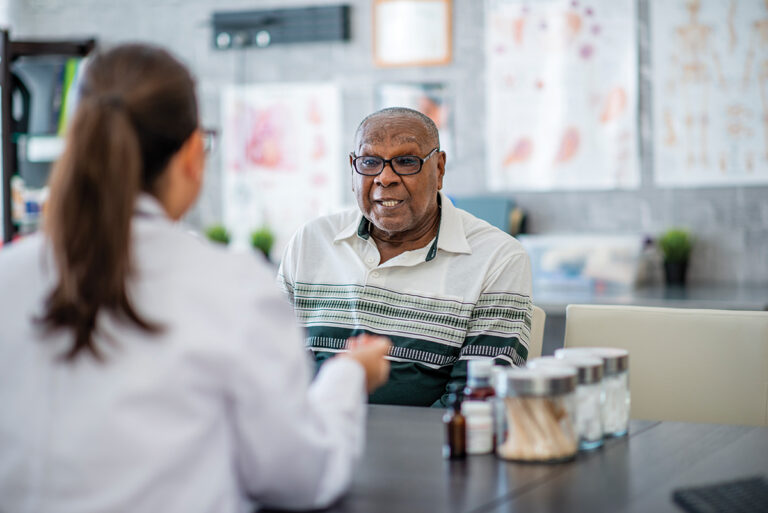 An elderly man looks at a pill bottle in his hand while sitting across the table from a doctor in the doctor's office. He appears to be making a decision about it.