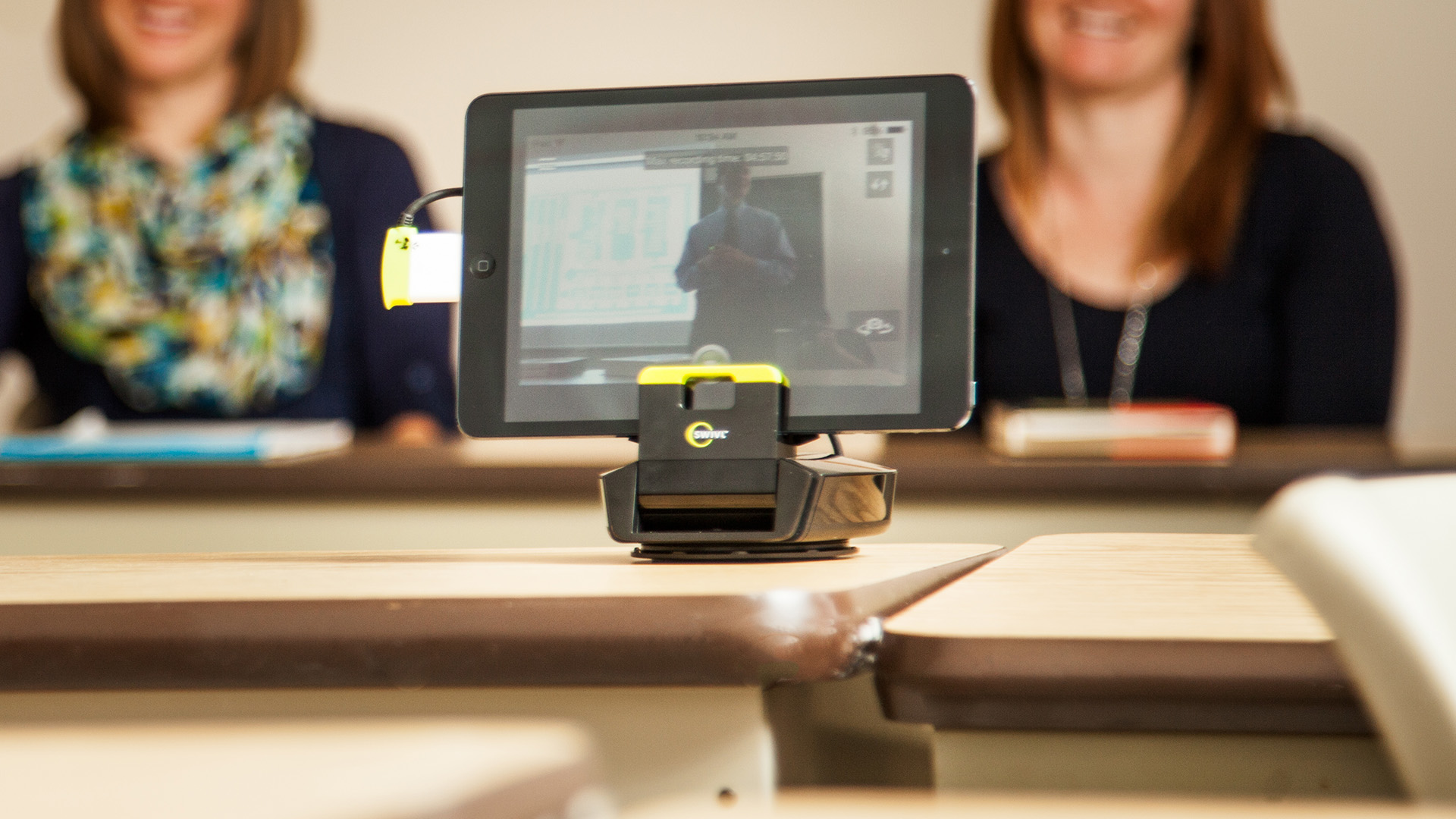 Two women watch a tablet where a teacher is presenting online