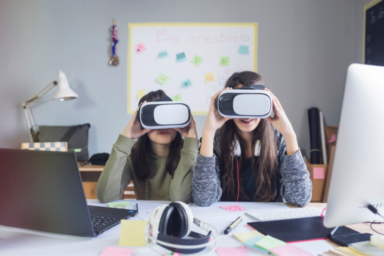 Two girls hold up VR sets near their computers at a work desk