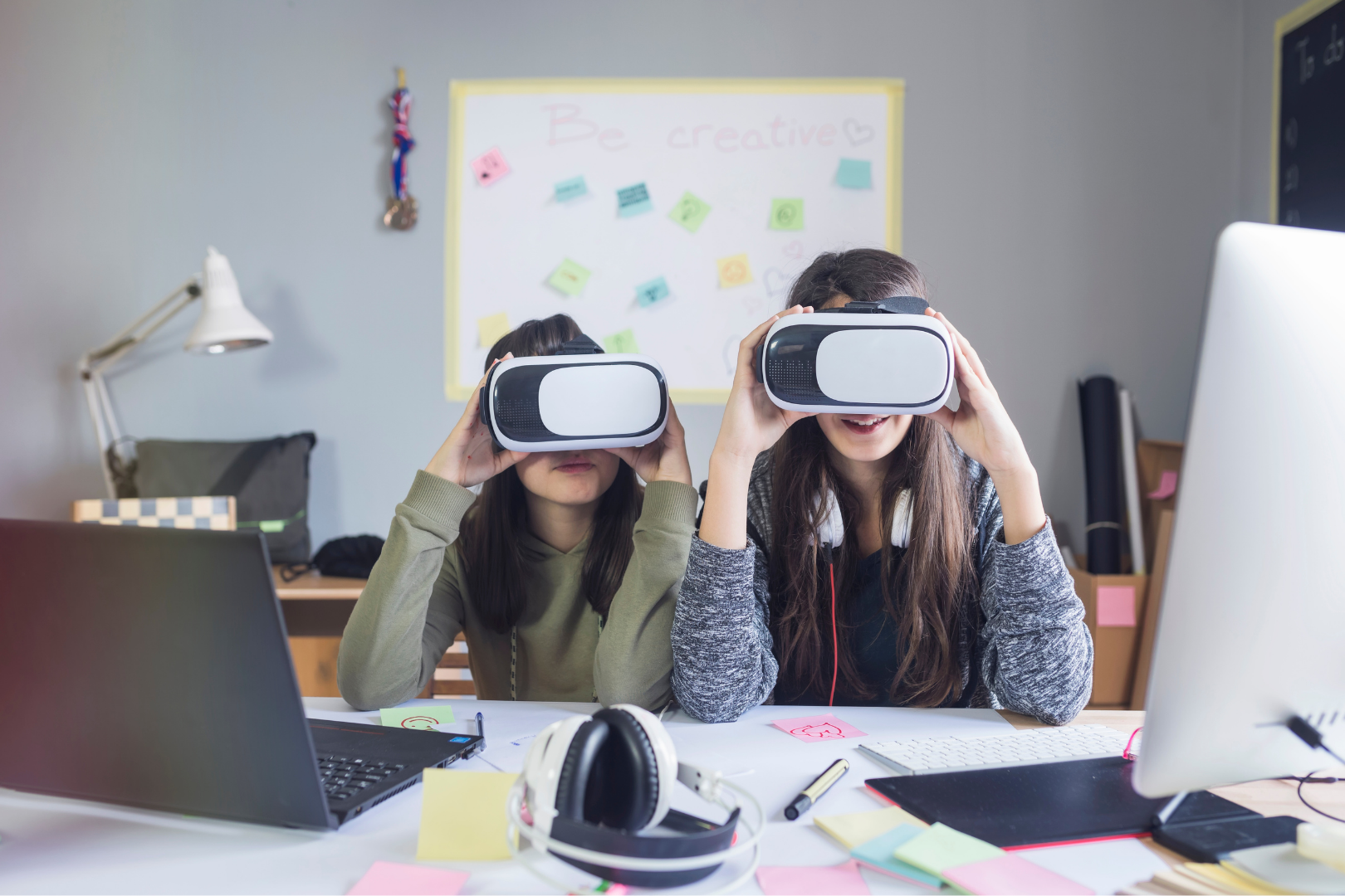 Two girls hold up VR sets near their computers at a work desk