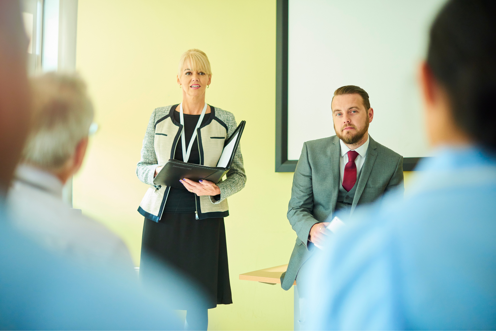 A legal nurse leads a meeting. She is dressed professionally and stands next a seated man in a suit. People in scrubs sit in the audience.