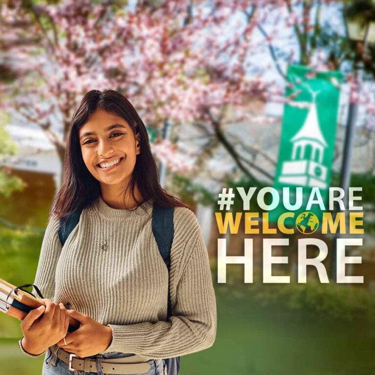 Smiling student with books and a backpack on the Wilmington University campus, with cherry blossoms and a banner in the background. Text reads “#YOU ARE WELCOME HERE.”
