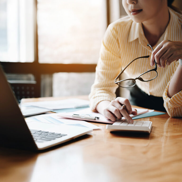 A professional in a yellow striped shirt works at a wooden desk, using a calculator and looking at a laptop. Documents and charts are spread out in front of them, suggesting a finance or accounting setting.