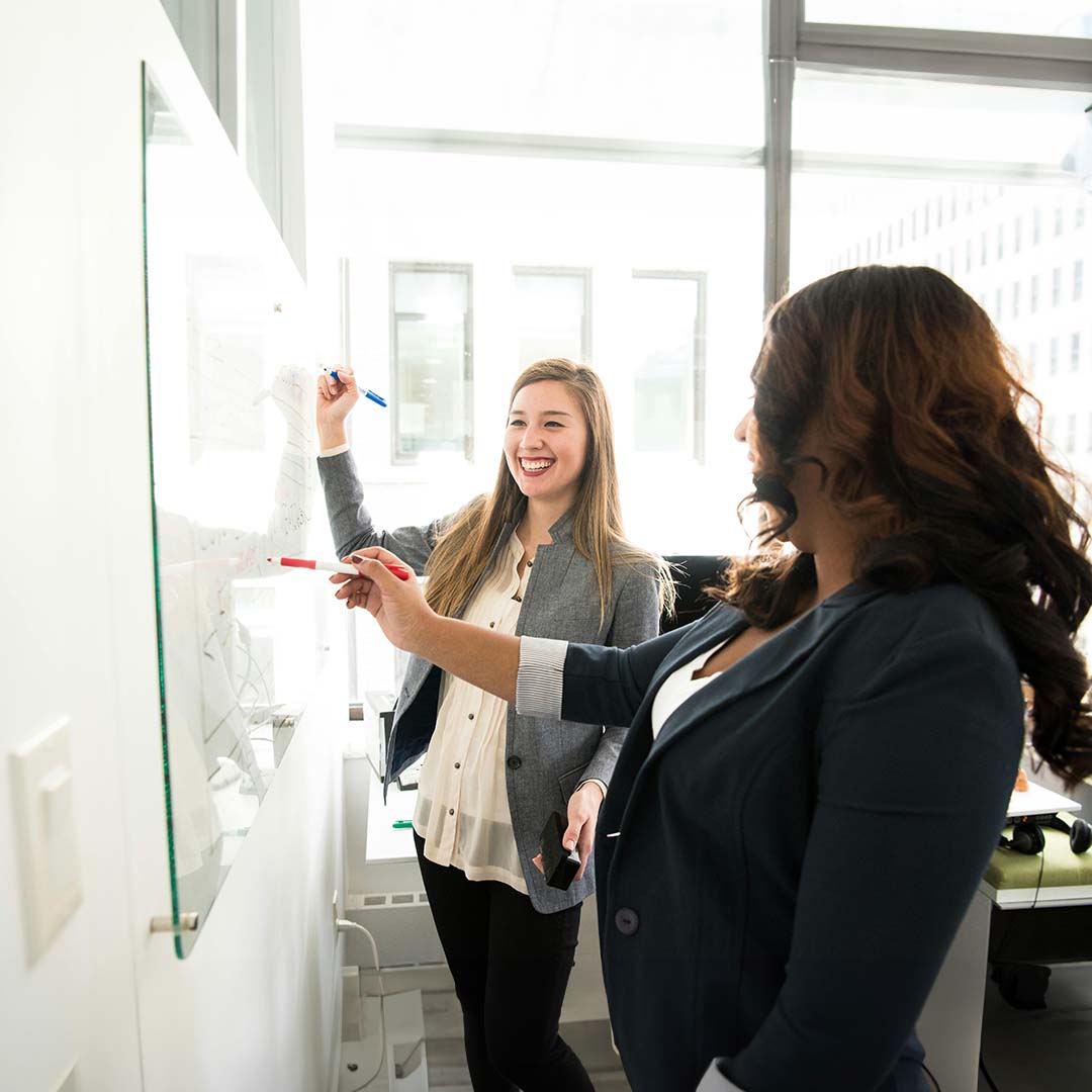 Two women in business attire collaborate at a whiteboard in a brightly lit office. One holds a blue marker and smiles, while the other points with a red marker.