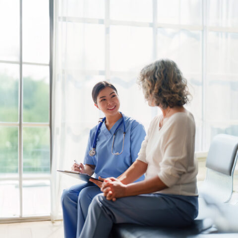 A smiling young woman in blue scrubs and a stethoscope sits on a bench, facing and speaking with an older woman with curly gray hair.