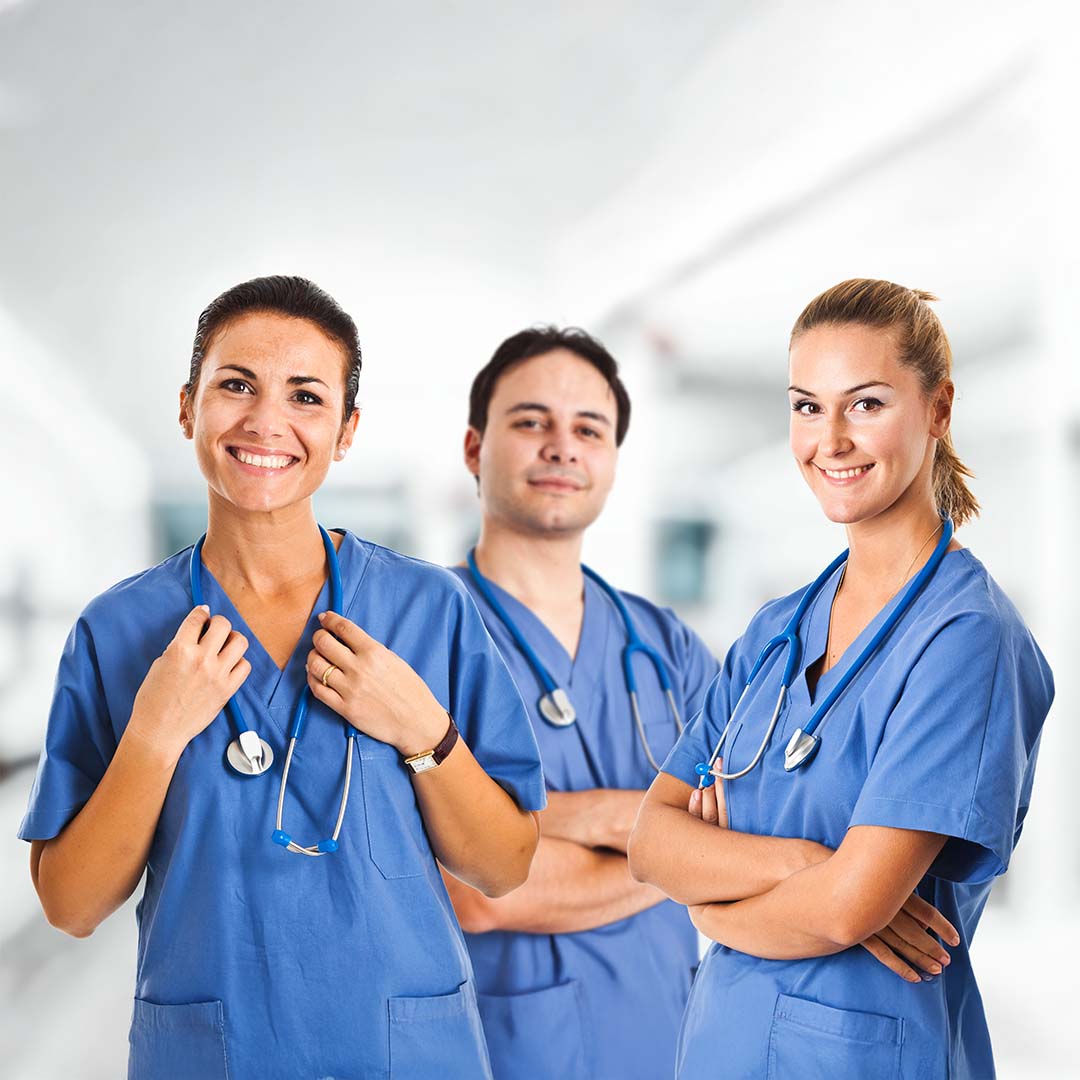 Three smiling healthcare workers in blue scrubs and stethoscopes stand in front of a blurred hospital hallway.