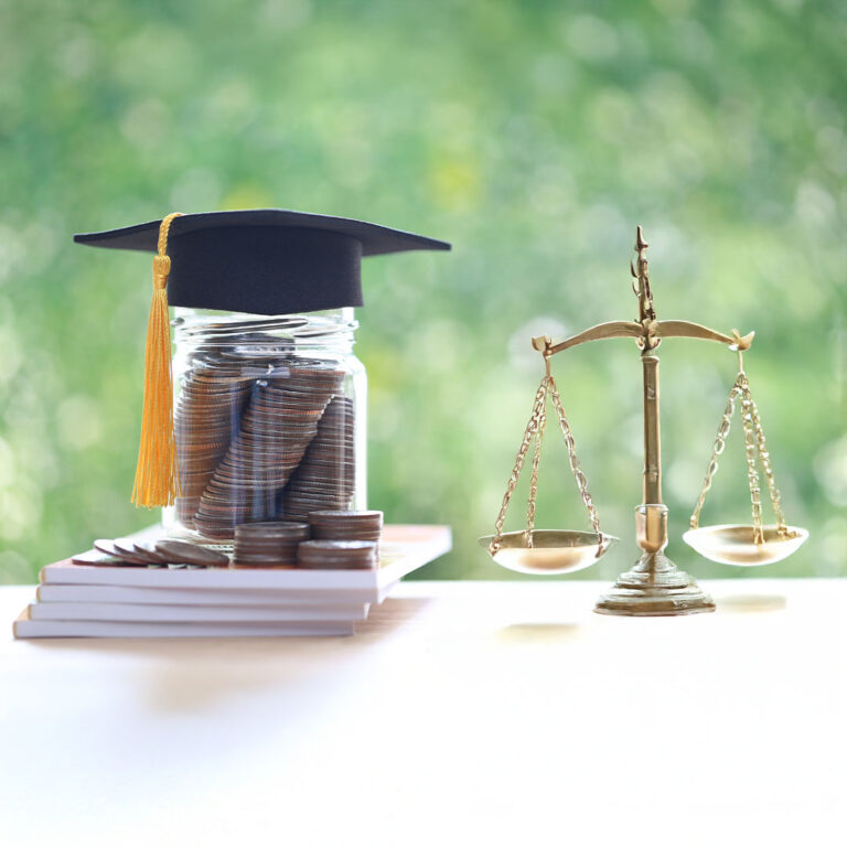 A jar of coins with a graduation cap on top next to a law scale on a table in front of a window.