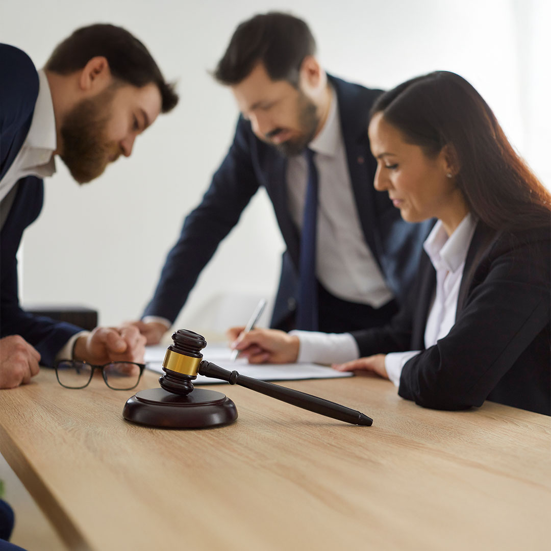 Three professionals in business attire are gathered around a light wooden table, seemingly reviewing documents or contracts. A wooden gavel with a gold band rests prominently in the foreground, suggesting a legal or judicial context.
