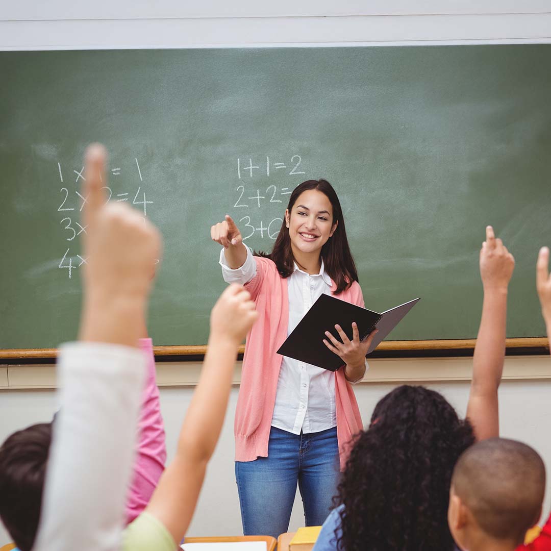 A teacher apprentice standing in front of a chalkboard and calling on one of their students thats raising their hand in front of them.