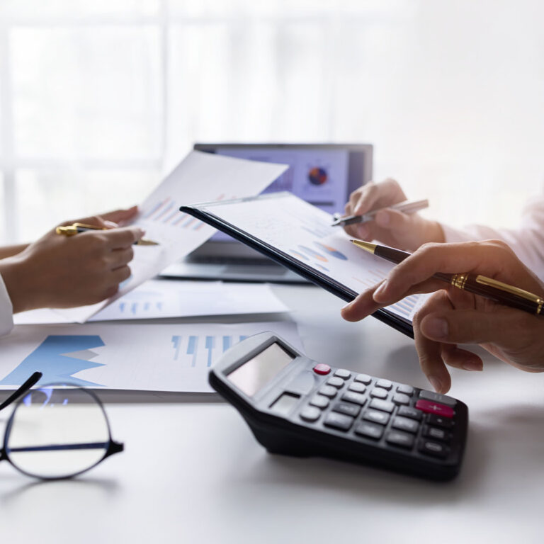 Two people are reviewing documents with charts and graphs on a table, which also holds a calculator and eyeglasses. The person on the right is pointing at a chart on a tablet, while the person on the left is holding up a paper with a chart.