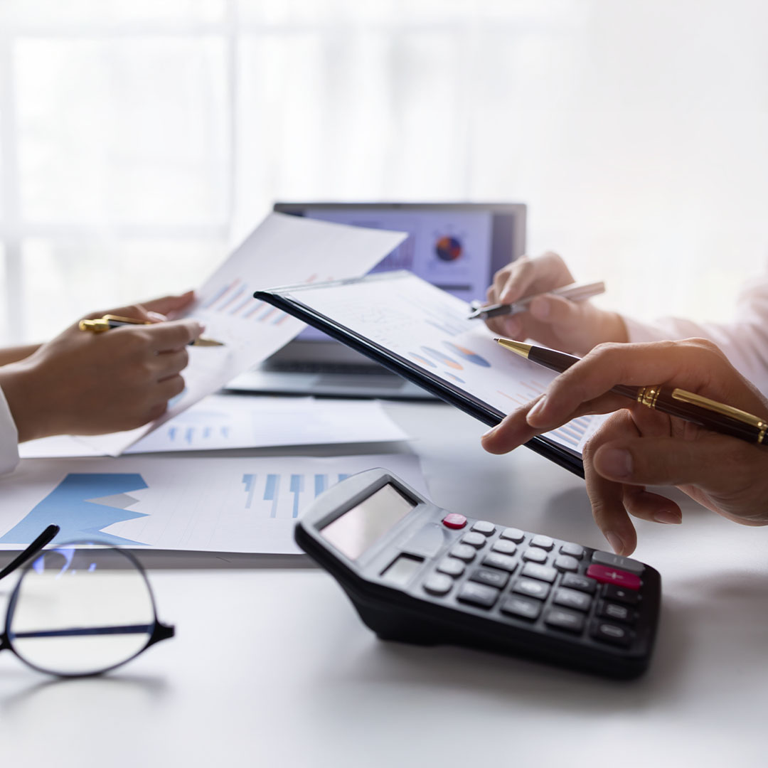 Two people are reviewing documents with charts and graphs on a table, which also holds a calculator and eyeglasses. The person on the right is pointing at a chart on a tablet, while the person on the left is holding up a paper with a chart.