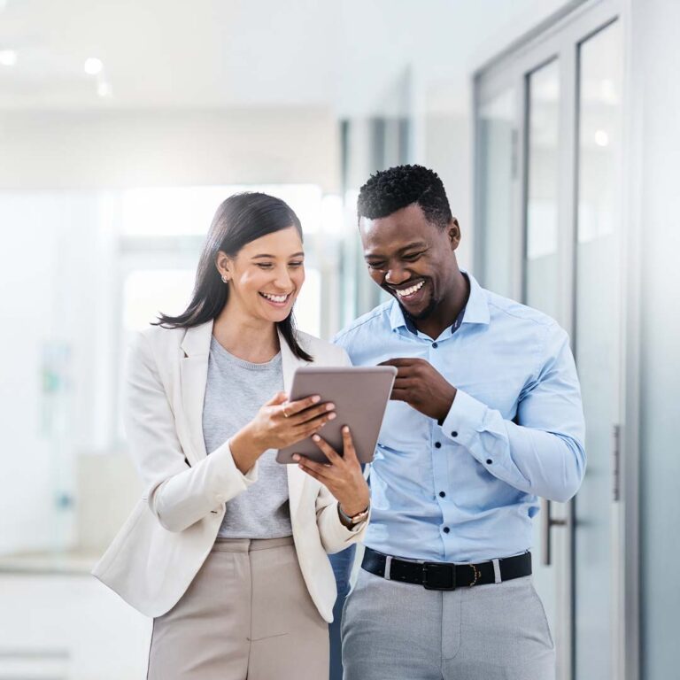 A smiling man in a light blue suit and a smiling woman in a white blazer and tan skirt stand together, looking at a tablet the woman is holding.