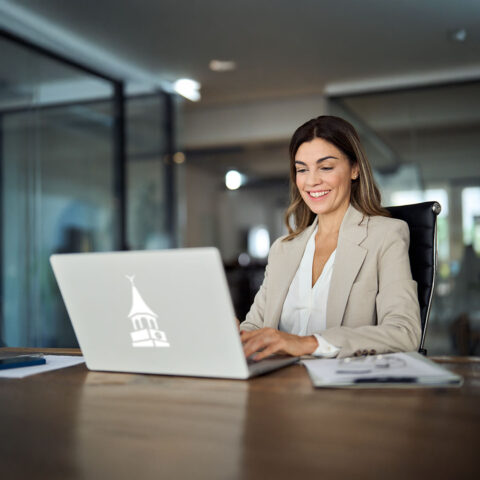 A smiling professional woman in a beige blazer sits at a wooden desk, working on a silver laptop. The laptop has a white WhilmU clocktower logo on the front.