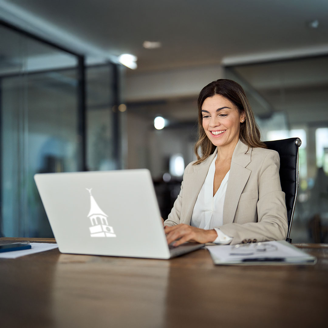 A smiling professional woman in a beige blazer sits at a wooden desk, working on a silver laptop. The laptop has a white WhilmU clocktower logo on the front.
