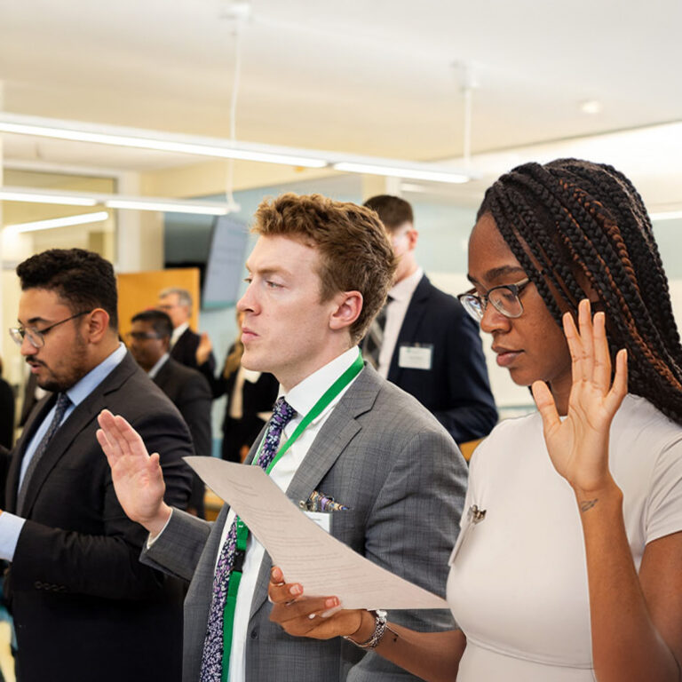 Three individuals in professional attire stand with their right hands raised, reciting an oath. A woman with braided hair, wearing glasses, is in the foreground on the right, holding a document.