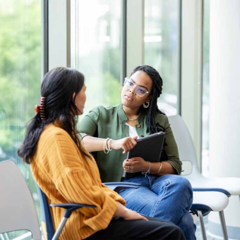 A professional woman with long braids and glasses sits in a bright office, holding a tablet and listening intently to a woman in a yellow sweater. The focus is on their supportive, face-to-face interaction.