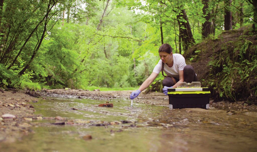 A female scientist sitting near the creek takes a sample of water.