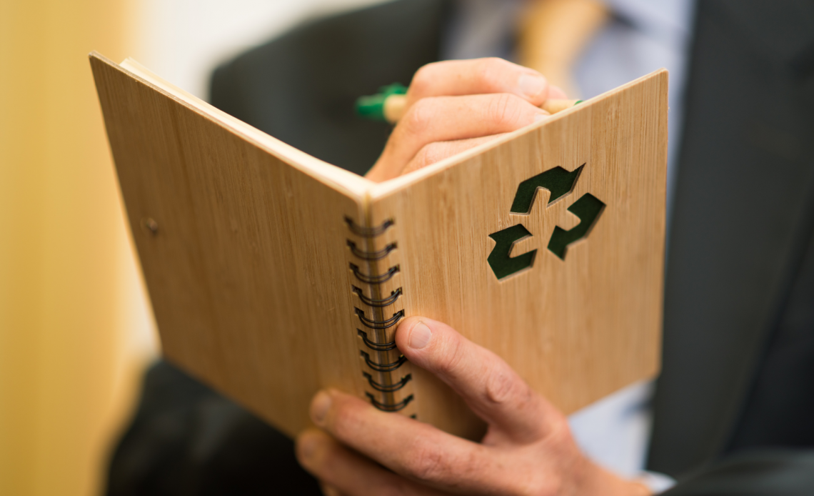 Close-up of a man's hands writing in a wooden notebook with a recycle symbol