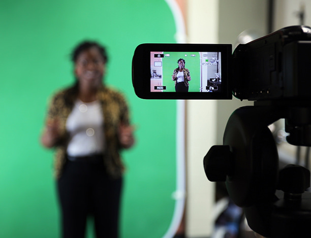 A woman speaks on a video camera screen. She stands in front of a green screen.