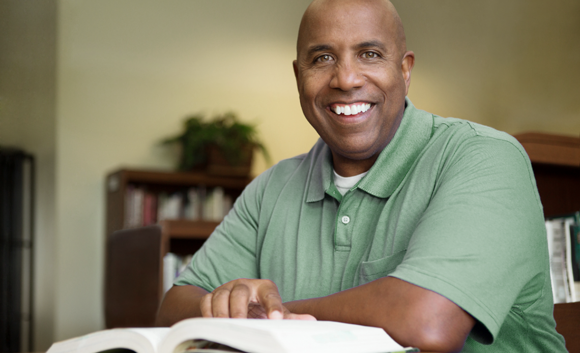 Adult student in green polo shirt smiles at camera while reading textbook
