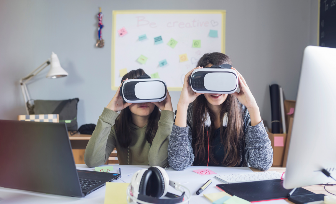 Two girls hold up VR sets near their computers at a work desk