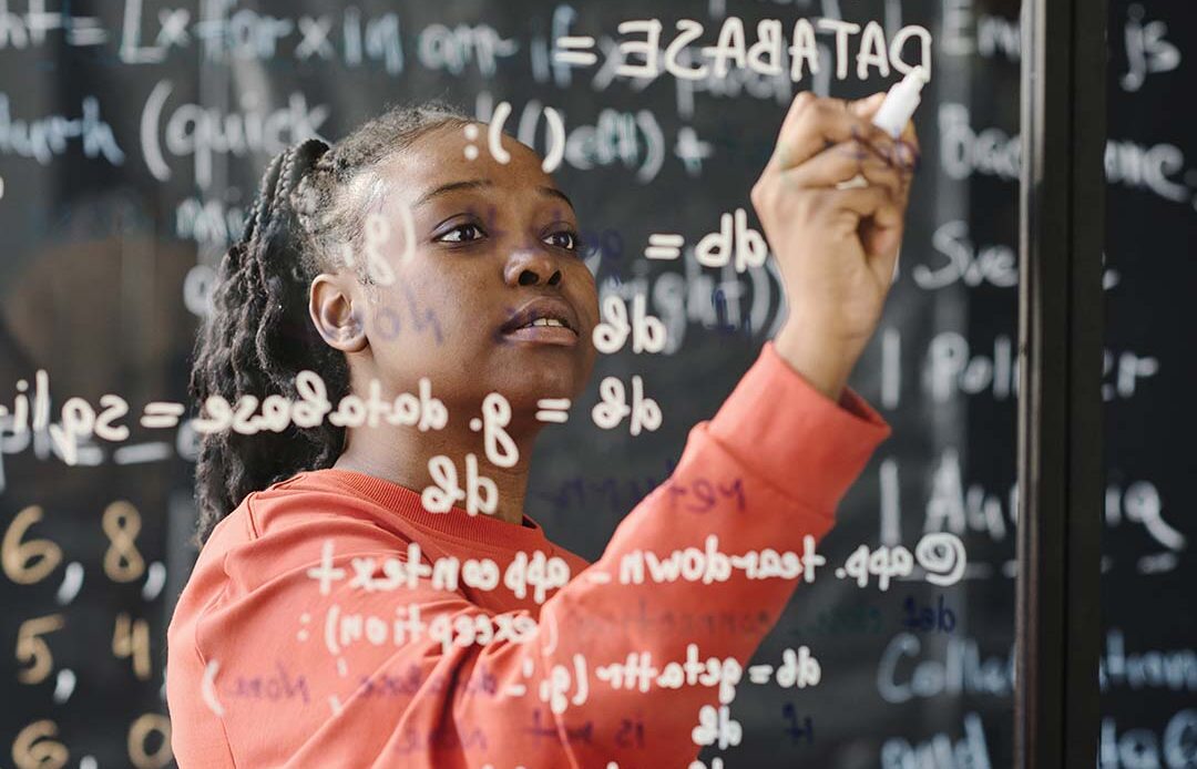 Student writing math equations on a clear board in a classroom.