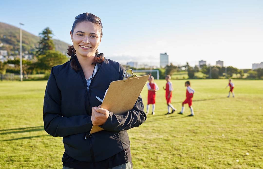 A smiling female coach holding a clipboard stands on a grassy soccer field with children in red uniforms practicing in the background.