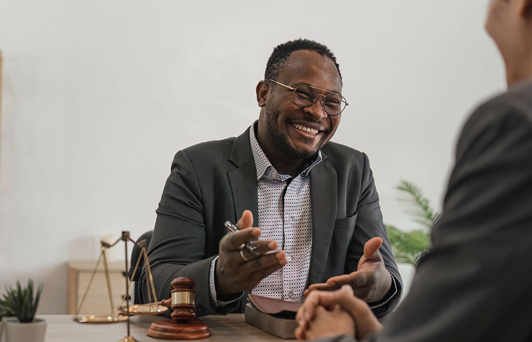 A man in a suit is sitting at a desk and smiling, engaged in conversation with another person across from him. He holds a pen in one hand while gesturing with the other. On the desk are legal items, including a gavel and scales of justice, suggesting a professional or legal setting. The background is minimal, with a plant and a small shelf in view.