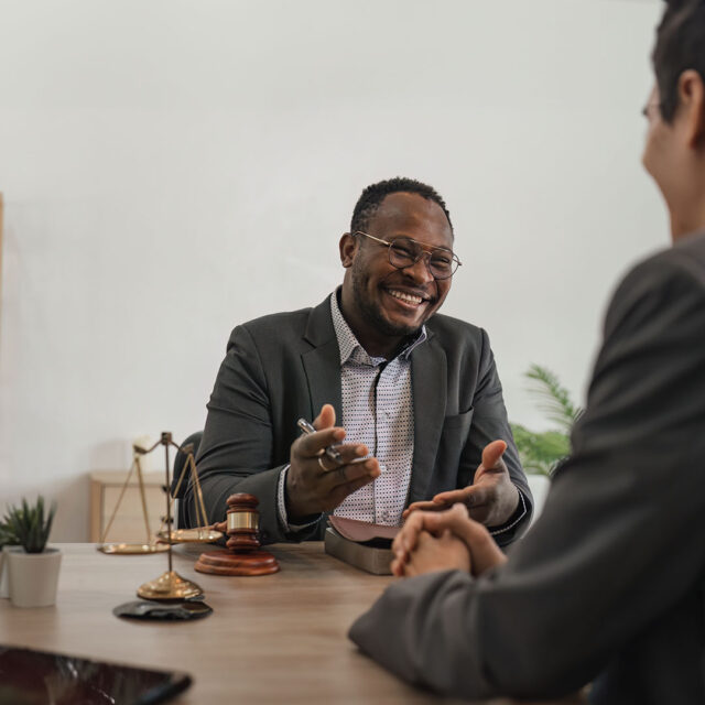 A man in a suit is sitting at a desk and smiling, engaged in conversation with another person across from him. He holds a pen in one hand while gesturing with the other. On the desk are legal items, including a gavel and scales of justice, suggesting a professional or legal setting. The background is minimal, with a plant and a small shelf in view.