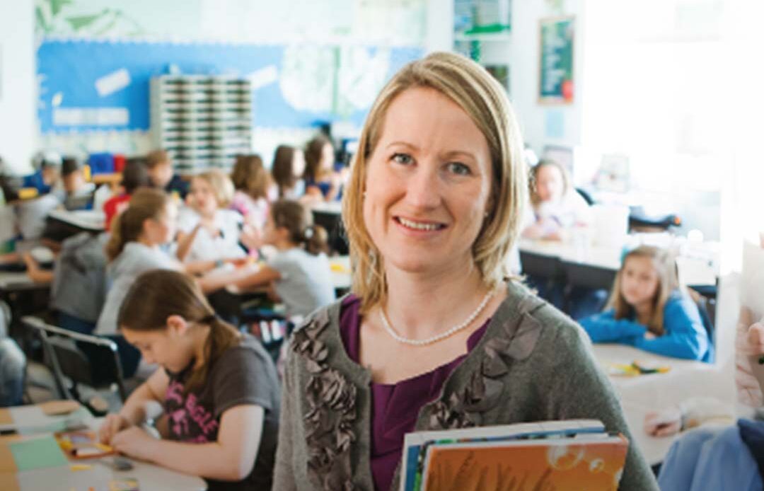 A smiling teacher holding a colorful picture book stands in a busy classroom with students working at desks in the background.